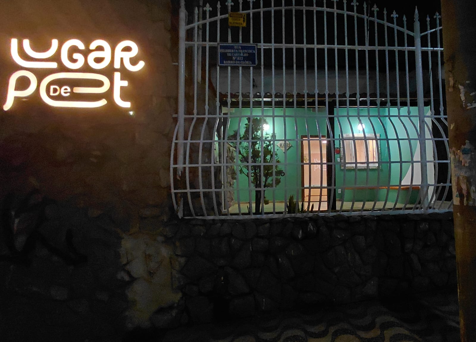 Fachada da Clínica, a clínica é uma casa de esquina com muro de pedra e portão de pedrestre e carro em grade branca. Em destaque aparece um letreiro luminoso com o nome da clínica: Lugar de Pet