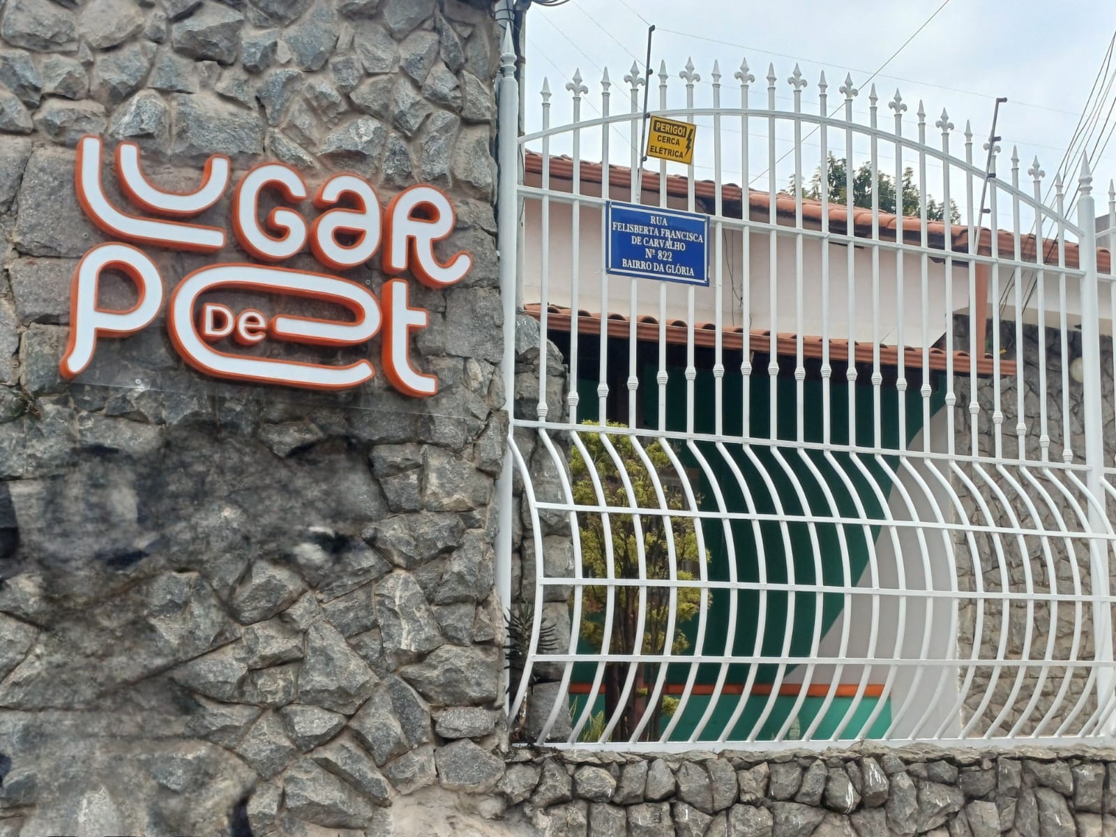 Fachada da Clínica, a clínica é uma casa de esquina com muro de pedra e portão de pedrestre e carro em grade branca. Em destaque aparece um letreiro luminoso com o nome da clínica: Lugar de Pet